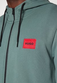 Mikina s kapucí v tlumené zelené barvě se zipem na přední straně. Na levé straně hrudi je červená čtvercová nášivka s logem "HUGO", černé stahovací šňůrky.