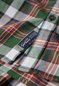 Geruit overhemd in groene, witte en oranje tinten; voorzien van een knoopsluiting, een gestructureerde stof en een blauw merkembleem bij de kraag.