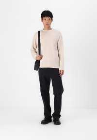Chemise beige à manches longues, pantalon noir et chaussures noires. Le mannequin tient un petit sac à bandoulière noir. Design minimaliste sur fond blanc.