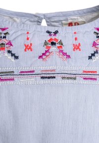 Valkoinen puuvillapaita, jossa on pystysuoria sinisiä raitoja. Kaula-aukon ympärillä on brodeeratut kukkakuviot vaaleanpunaisena, oranssina, vihreänä ja mustana.