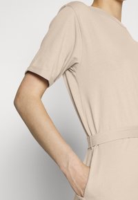 Vestido bege com mangas curtas, tecido suave, corte solto e cintura cingida. Apresenta bolsos laterais e decote redondo.