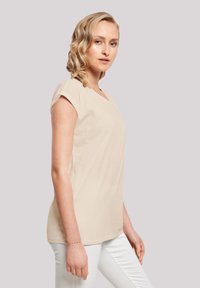 Beige short-sleeve t-shirt with a v-neck design, made from soft fabric. The model pairs it with slim white trousers, highlighting a casual look.