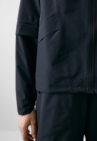 Navy jacket featuring a zip closure, multiple pockets, and a drawstring hem. Made from a textured, water-resistant material.