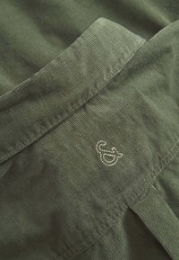 Tessuto di velluto a coste verde oliva con una texture a coste. Presenta un piccolo emblema decorativo argentato sul colletto. Cuciture ordinate visibili.