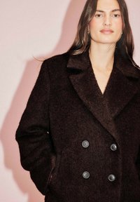 Dvigubas tamsiai rudas paltas su tekstūruotu audiniu, dideliais atvartais ir dvigubu sagčių uždarymu. Subtilus blizgesys ir pritaikyta siluetas.