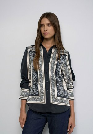 Mujer con camisa negra abotonada con patrones florales ornamentados en blanco y azul, de pie con los brazos relajados a los lados frente a un fondo liso.