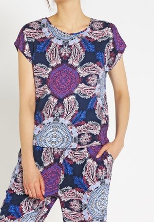 Vrouw draagt een bijpassende korte mouw top en broek met een kleurrijk ingewikkeld paisleymotief in blauwtinten, paars en wit.