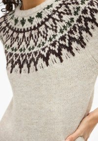 Zbliżenie beżowego swetra dzianego z ciemnobrązowymi i zielonymi geometrycznymi wzorami wokół dekoltu, noszonego przez osobę z kręconymi włosami.