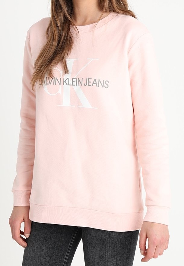 Femme portant un sweat-shirt Calvin Klein Jeans rose clair avec des lettres de logo blanches et grises, associé à un jean gris foncé.