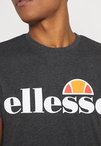 Camiseta de color gris oscuro con el logotipo blanco de "ellesse". Presenta un gráfico en forma de medio círculo en colores naranja, amarillo y rojo por encima de la letra.