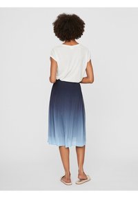 Vrouw met krullend haar, gekleed in een witte top met korte mouwen, een blauw ombre geplooide midi-jurk en beige instap-schoenen, staand met haar rug naar de camera.