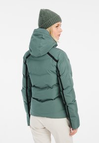 Chaqueta acolchada verde con capucha, que presenta secciones acolchadas y acentos laterales negros. El modelo lleva un gorro de punto verde texturizado.