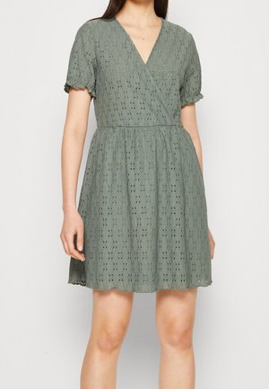 Kobieta w oliwkowo-zielonej, krótkorękawowej sukience kopertowej z fakturą, z falistymi krawędziami i wzorem z dziurek, stojąca na jednolitym tle.