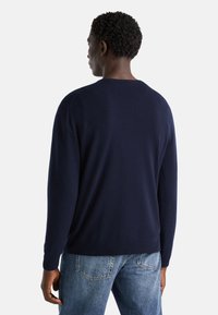 Granatowy sweter z wełny z domieszką z długim rękawem, okrągłym dekoltem, ściągaczami na rękawach oraz luźnym krojem, w zestawieniu z jasnoniebieskimi dżinsami.