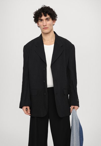 Černý oversized blazer s výstřihem do "V", jednoprocentovými knoflíky a dvěma předními kapsami, oblečený přes bílou košili. Modrá džínsová bunda v ruce.