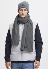 Grå strikket hue og tørklæde sat mod en lys grå puffervest og marineblå sweatshirt. Struktureret stof med mærkeetiket på tørklædet.