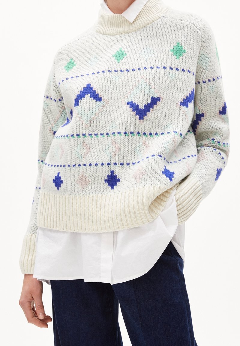 Personne portant un pull en maille crème avec des motifs géométriques verts et bleus sur une chemise blanche et un pantalon bleu foncé, main dans la poche.