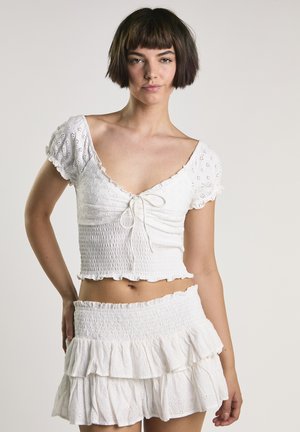 Ung kvinde med kort mørkt hår iført en hvid crop top med hulmønster og puffede ærmer samt et mini-skørt med lagdelte flæser.