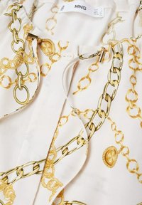 Tissu blanc avec motif chaîne et médaillon dorés, avec un cordon à embouts métalliques dorés près de l’étiquette MNG.