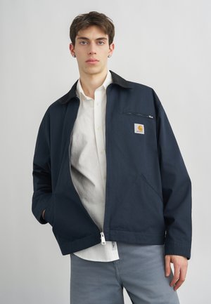 Fiatal férfi sötétkék Carhartt cipzáras dzsekiben, fehér gombos inggel és világosszürke nadrággal, sima háttér előtt állva.