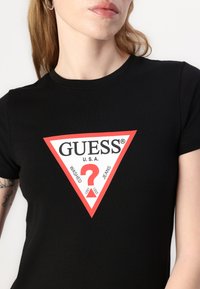 T-shirt en coton noir avec un graphique triangulaire rouge portant les inscriptions « GUESS® », « U.S.A. », « WASHED » et un point d'interrogation rouge. Manches courtes et col rond.