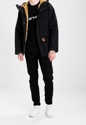 Mężczyzna w czarnej kurtce z beżowym futerkiem, czarnych spodniach, czarnej koszulce Carhartt, białych sneakersach oraz z widocznymi tatuażami na rękach, stojący na jednolitym tle.