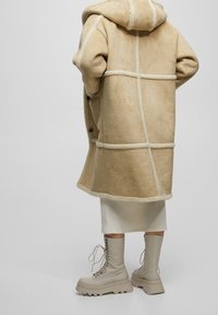 Beige shearling frakke med hætte, der har et rutemønster og blød tekstur. Båret over en ribbet hvid nederdel og lyse snørebåndsstøvler.