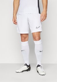 Pantaloni corti da calcio Nike bianchi con logo nero, abbinati a calze bianche fino al ginocchio con un accento nero. Scarpini da calcio bianchi e neri.
