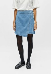 Personne portant une jupe portefeuille en denim bleu, des collants noirs, des mocassins noirs et une chemise blanche à manches courtes, debout devant un arrière-plan uni.