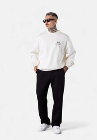 White sweatshirt with logo, black pants, and white sneakers; model also wearing black sunglasses; casual fit with dropped shoulders.