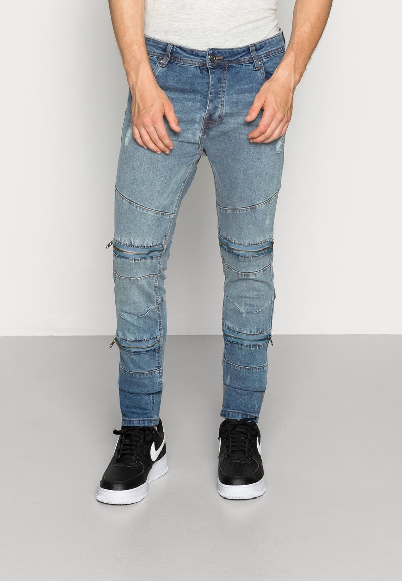 Ljusblå denimjeans med slim fit, med flera dragkedjedetaljer och paneler, som bärs med svarta sneakers på en neutral bakgrund.