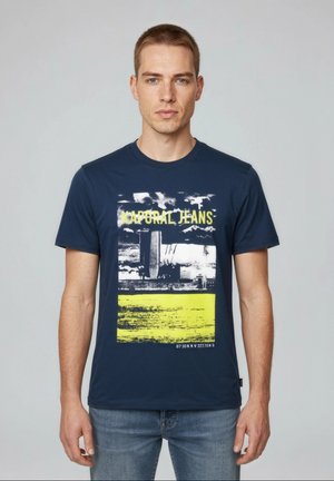 Junger Mann mit kurzen Haaren, der ein dunkelblaues Kaporal Jeans T-Shirt mit einer Stadtansicht und gelbem Grafikdruck trägt, kombiniert mit hellblauen Jeans.