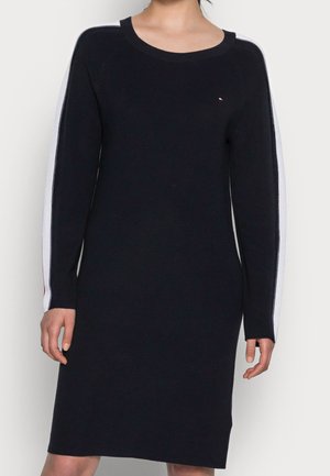 Zwarte gebreide jurk met lange mouwen, witte strepen op de mouwen en een klein rood-wit-blauw logo op de linkerborst.