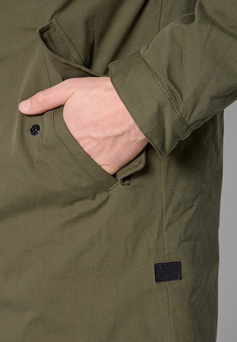 Chaqueta verde oliva con tela texturizada, cierre de botones a presión, gran bolsillo lateral y una etiqueta de marca negra en la costura inferior.