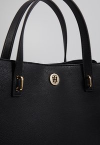 Sac fourre-tout en cuir noir texturé avec double poignées, présentant un accent logo doré et des attaches en boucle sur les côtés.