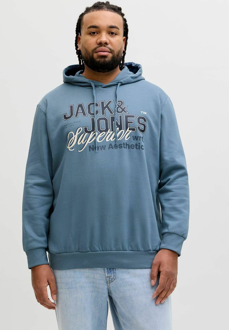 Mann mit geflochtenem Haar trägt einen blauen Jack & Jones Hoodie und hellblaue Jeans, steht vor einem schlichten weißen Hintergrund.