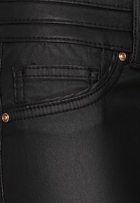 Jeans de mezclilla negra con una textura suave, que presentan un pequeño bolsillo frontal, costuras prominentes y acentos de herrajes en color cobre.