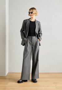 Blazer gris à motifs pied-de-poule avec un top noir, un pantalon gris plissé taille haute et des chaussures noires imposantes. Mannequin portant des lunettes de soleil.