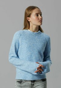 Młoda kobieta w jasnoniebieskim wełnianym swetrze z długimi rękawami, skrzyżowane ręce, zwrócona w prawo na jednolitym szarym tle.