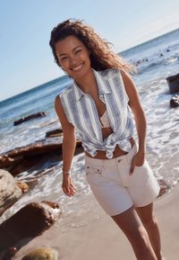 Chemise sans manches à rayures bleu clair et blanc nouée à la taille, associée à un short blanc, portée près de la plage avec un littoral rocheux.