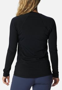 Frau mit blonden Haaren, die ein enges schwarzes Langarmshirt und blaue Leggings trägt, von hinten vor einem schlichten weißen Hintergrund gezeigt.