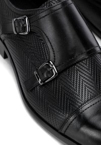 Zapato de vestir negro de cuero con patrón de espiga, acentos de doble hebilla, textura suave y punta brillante.