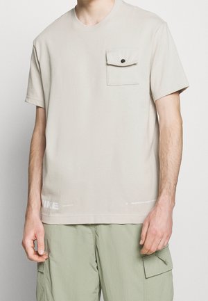 Homme portant un t-shirt à manches courtes beige clair avec une poche poitrine boutonnée et un pantalon cargo vert clair.