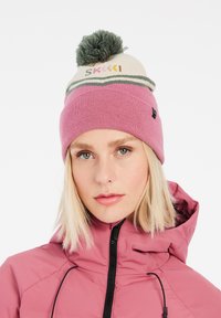 Gehaakte beanie in zachtroze, met een crème band voorzien van de tekst "Ski", afgewerkt met een groene pompon en smalle strepen.
