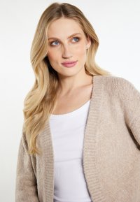 Beige gebreide cardigan met een gestructureerde afwerking, open voorkant en geribde randen, gedragen over een witte aangesloten top.