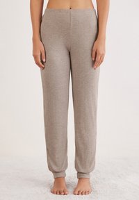 Femme portant un pantalon de lounge en tissu doux beige, debout pieds nus sur un tapis clair devant un fond uni.
