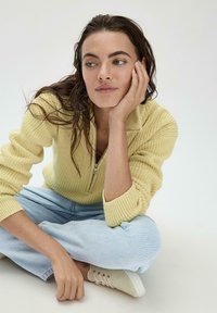 Gelber gestrickter Pullover mit einem halben Reißverschluss-Kragen, gerippter Textur und langen Ärmeln, kombiniert mit hellblauen Jeans und beigen Schuhen.