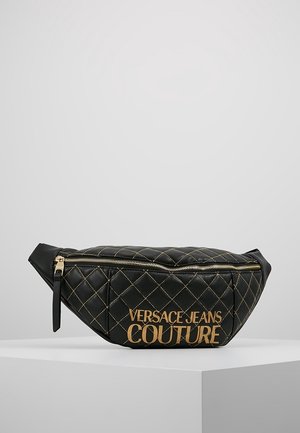 Zwarte doorgestikte leren Versace Jeans Couture heuptas met gouden stiksel en gouden rits op een wit voetstuk.