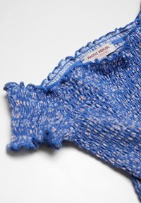 Haut bleu smocké avec bords froncés, présentant un tissu texturé avec des éclats multicolores. L'étiquette indique "PACIFIC REPUBLIC."