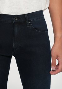 Jeans in denim scuro con un design aderente, stile a cinque tasche, cuciture a contrasto sottili e chiusura con bottone classico. Texture liscia.